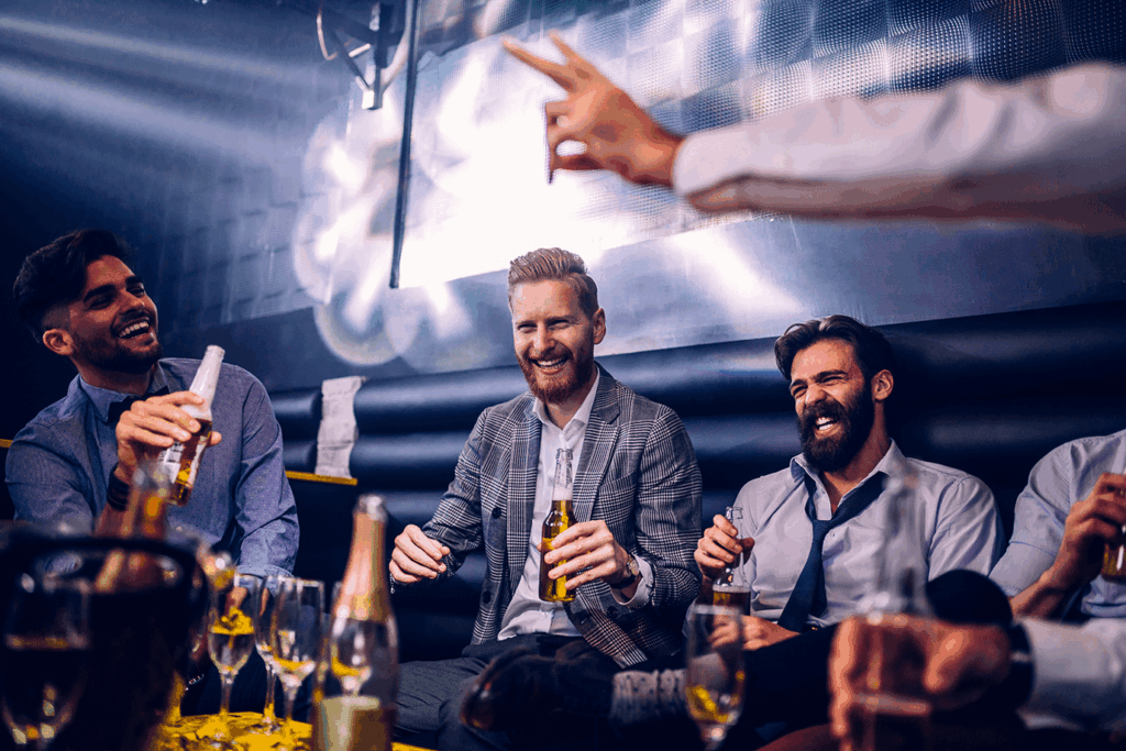 Trois hommes souriants et barbus rient en buvant de la bière lors d'une soirée animée dans un club. Des verres et bouteilles sont visibles sur la table.