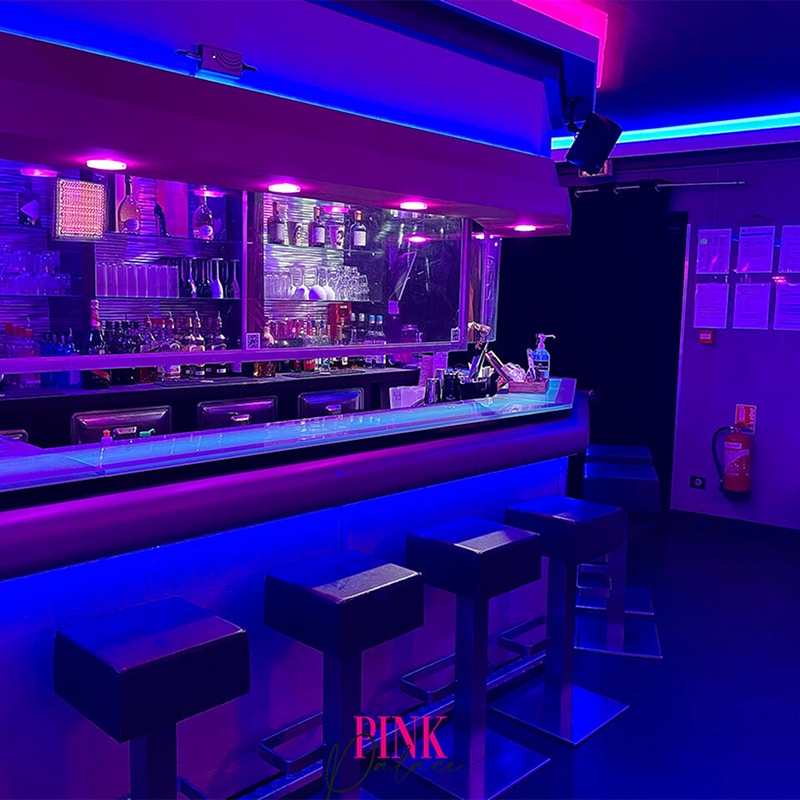 Bar moderne illuminé par des néons roses et bleus. Comptoir élégant avec tabourets, bouteilles et verres. Ambiance club/lounge "PINK".