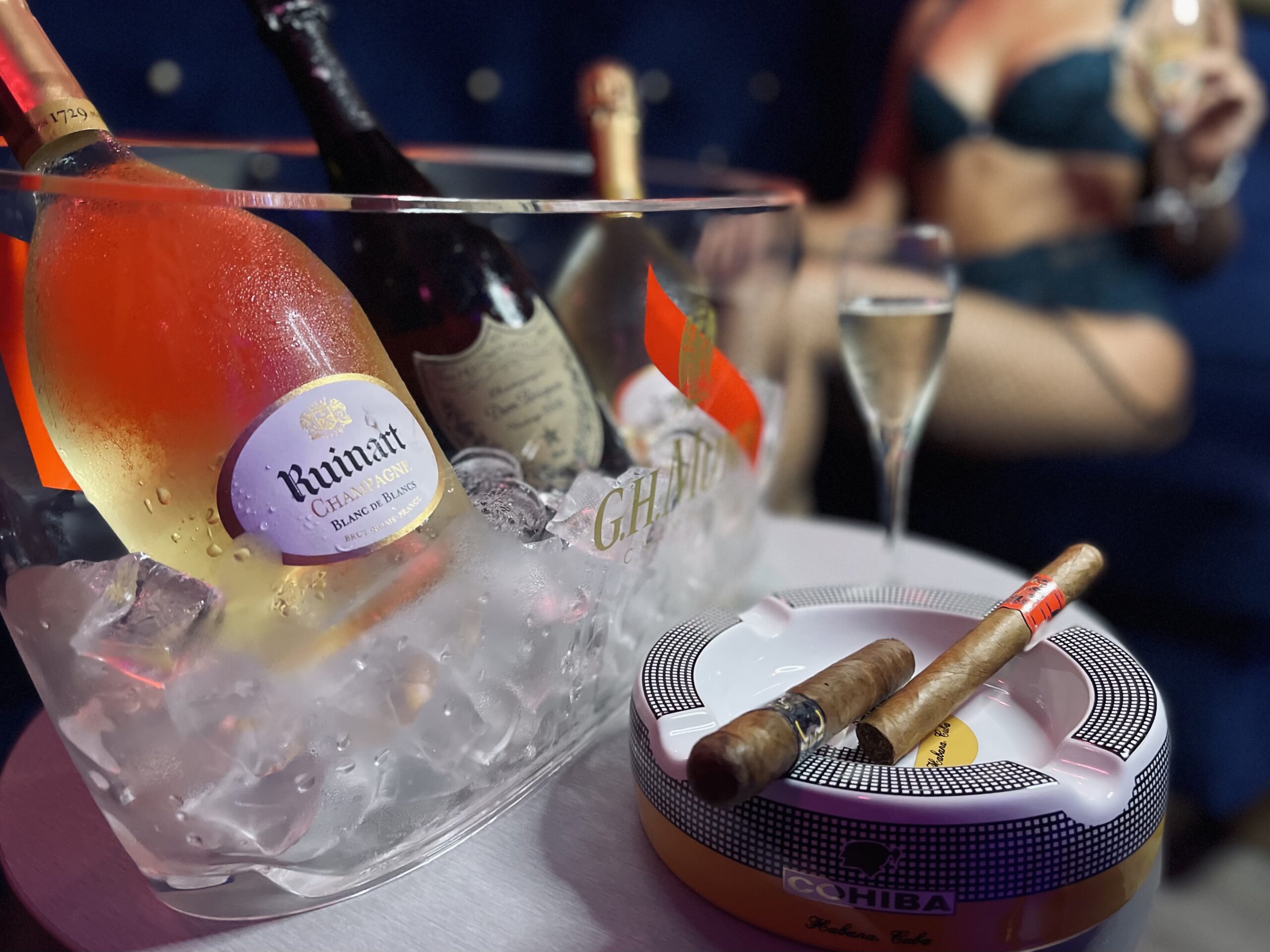 Ambiance luxueuse avec bouteille de champagne Ruinart dans un seau à glace, cigares Cohiba sur un cendrier, et femme élégante tenant un verre de bulles.