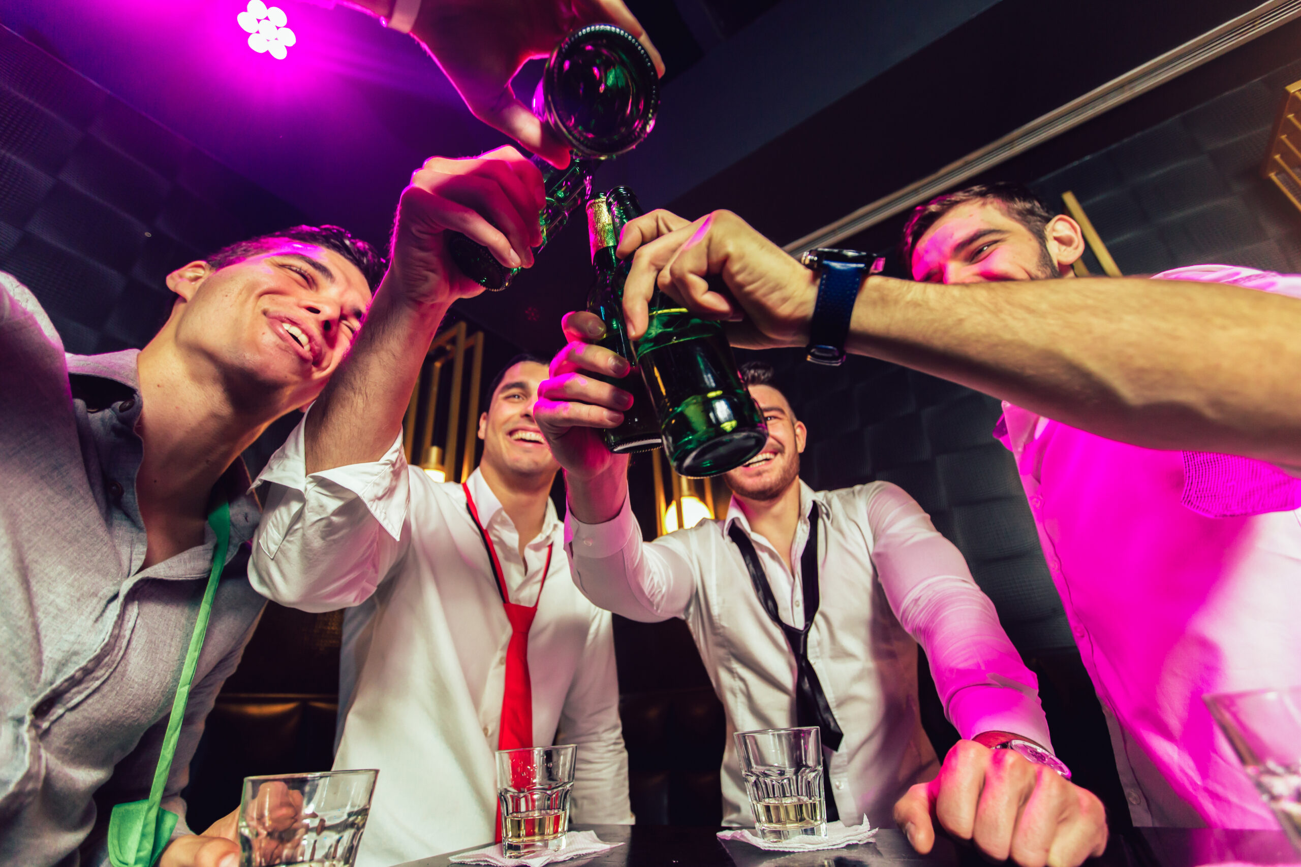 Quatre hommes joyeux trinquent des bières dans un bar animé, éclairé par des lumières néon violettes. Scène de fête entre amis.