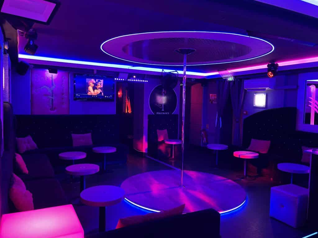 Club de pole dance néon bleu et rose. Scène centrale, canapés lounge, tables basses, TV, et enseigne 'POLE DANCE'.
