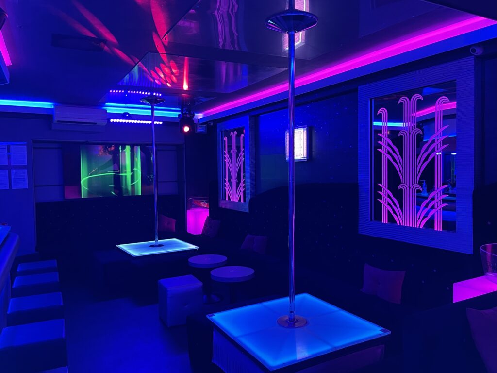 Club néon moderne avec deux pôles de danse lumineux bleus, banquettes sombres, éclairage rose et bleu, et miroirs Art Déco.
